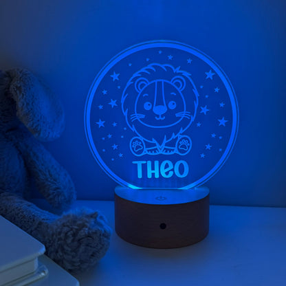 A personalized 3D optical illusion night light shaped like a circular wooden base with a glowing blue lion design and the name 'THEO' on the front, shown on a tabletop next to a teddy bear.