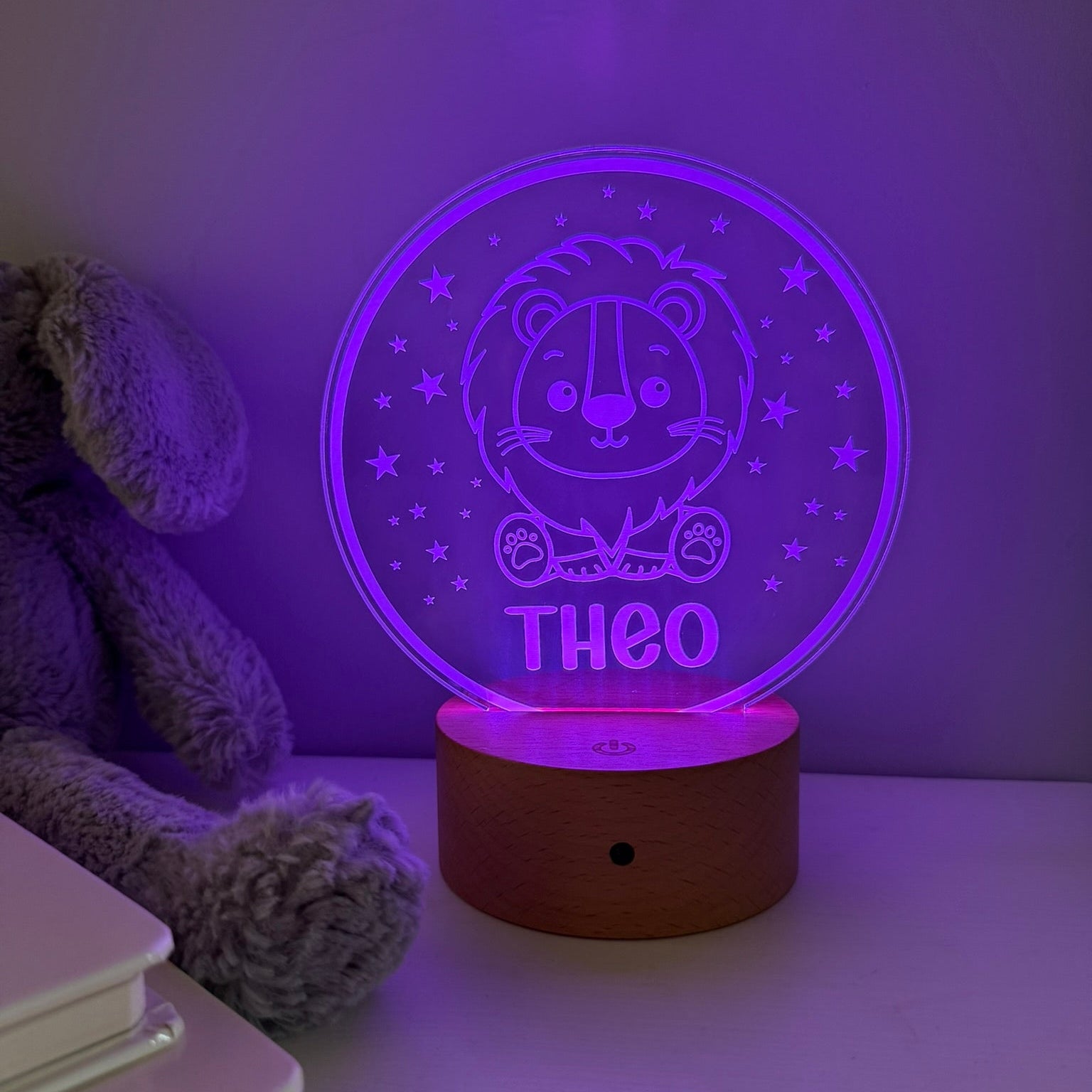 A personalized 3D optical illusion night light with a cartoon lion and the name 'THEO' on a glowing purple circular disc, resting on a wooden base on a tabletop next to a plush toy.