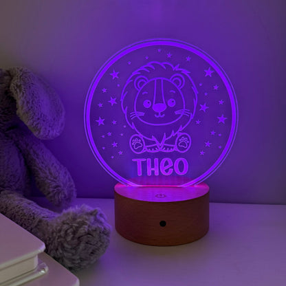 A personalized 3D optical illusion night light with a cartoon lion and the name 'THEO' on a glowing purple circular disc, resting on a wooden base on a tabletop next to a plush toy.