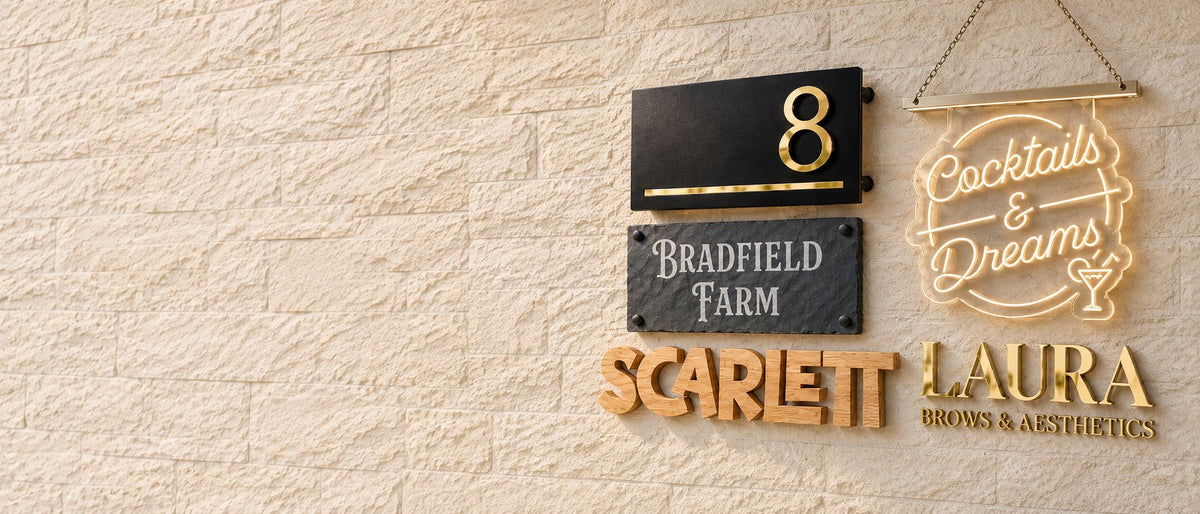 Luxe Sign Studio signs on a pale brick wall, including a house number 8 sign, a slate Bradfield Farm sign, a lit Cocktails & Dreams sign and wooden and gold name lettering