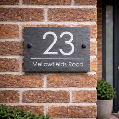 House number 23 Mellowfields Road on a dark slate plaque against a brick wall.