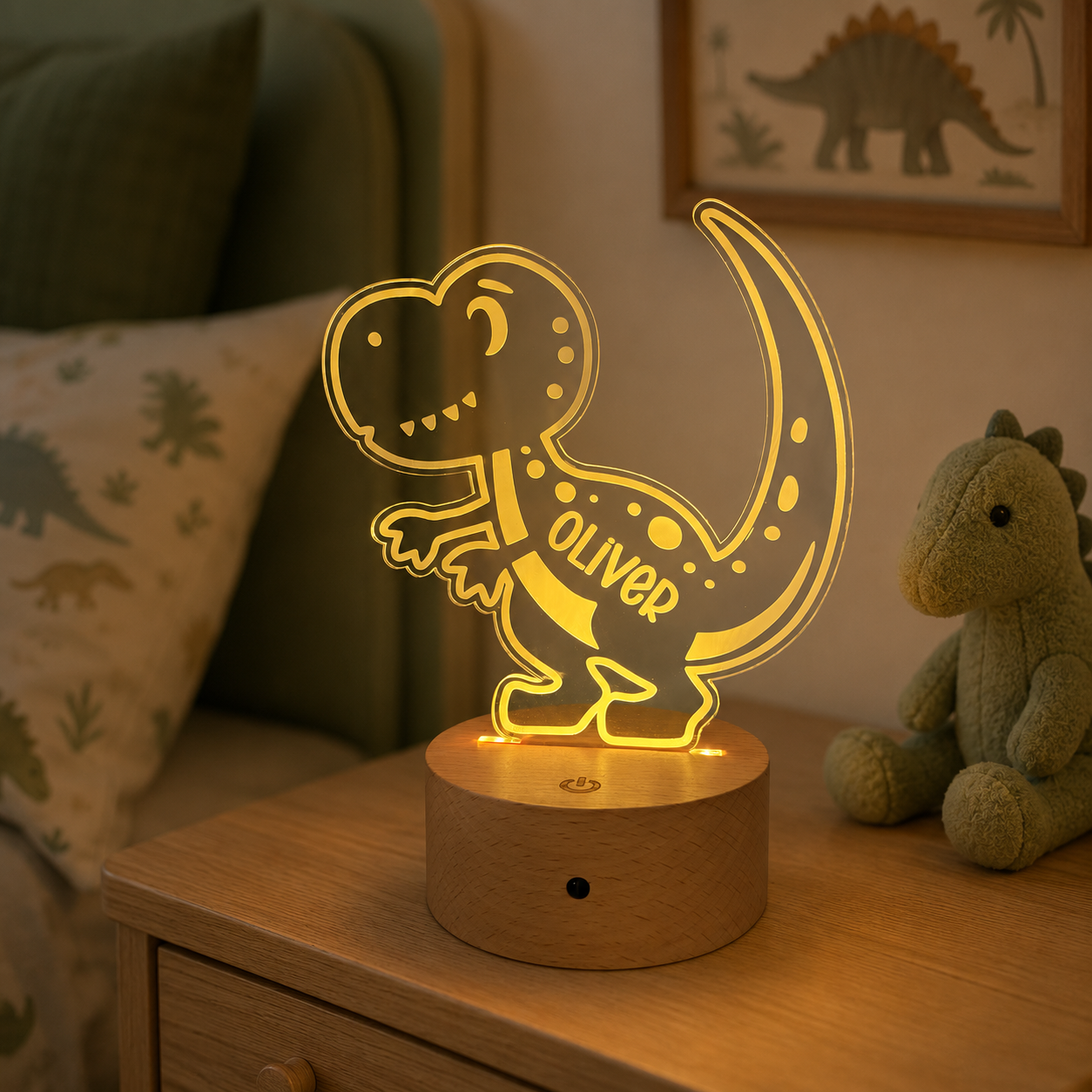 A personalized LED night light shaped like a cartoon T-rex dinosaur, glowing with warm yellow light on a round wooden base. The word 'OLIVER' is printed on the side of the dinosaur.